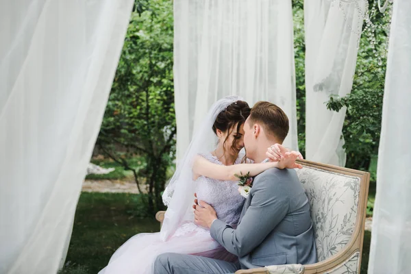 Wedding. The bride and groom are sitting on a beautiful couch in a gazebo in the garden. The bride in a pink dress, the groom in a gray suit. White tent. The bride and groom are walking in the forest.