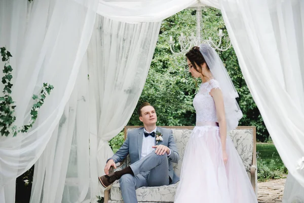 Boda. La novia y el novio están sentados en un hermoso sofá en un mirador en el jardín. La novia con un vestido rosa, el novio con un traje gris. Carpa blanca. La novia y el novio están caminando en el bosque . —  Fotos de Stock