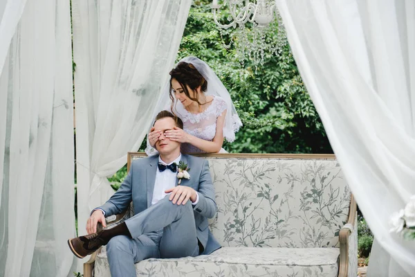 Hochzeit. Braut und Bräutigam sitzen auf einer schönen Couch in einem Pavillon im Garten. die Braut im rosa Kleid, der Bräutigam im grauen Anzug. Weißes Zelt. Braut und Bräutigam spazieren im Wald. — Stockfoto