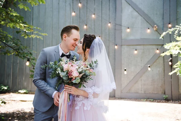 Hochzeit. Porträt einer schönen Braut und Bräutigam. Vor dem Hintergrund einer Girlande aus Glühbirnen und Metalltüren spazieren Braut und Bräutigam durch den Garten. Strauß von Grün und Blumen. — Stockfoto