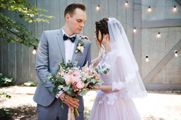 Hochzeit. Porträt einer schönen Braut und Bräutigam. Vor dem Hintergrund einer Girlande aus Glühbirnen und Metalltüren spazieren Braut und Bräutigam durch den Garten. Strauß von Grün und Blumen. — Stockfoto