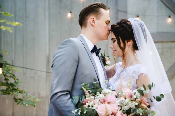 Hochzeit. Porträt einer schönen Braut und Bräutigam. Vor dem Hintergrund einer Girlande aus Glühbirnen und Metalltüren spazieren Braut und Bräutigam durch den Garten. Strauß von Grün und Blumen. — Stockfoto