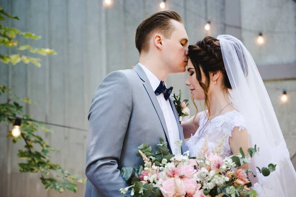 Hochzeit. Porträt einer schönen Braut und Bräutigam. Vor dem Hintergrund einer Girlande aus Glühbirnen und Metalltüren spazieren Braut und Bräutigam durch den Garten. Strauß von Grün und Blumen. — Stockfoto