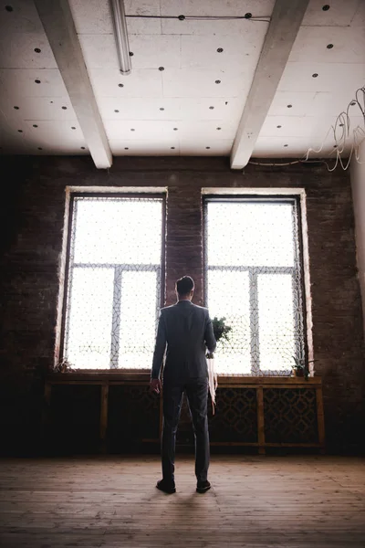 El novio está esperando a la novia con un traje gris. Novio sosteniendo ramo y esperando a la novia. Día de la boda. El novio está cerca de una gran ventana . — Foto de Stock