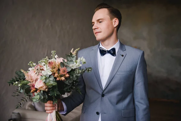 Lo sposo sta aspettando la sposa con un completo grigio. Sposo in possesso di bouquet e in attesa di sposa. Il giorno del matrimonio. Lo sposo si trova vicino a una grande finestra . — Foto Stock