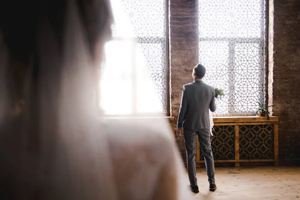 El novio está esperando a la novia con un traje gris. Novio sosteniendo ramo y esperando a la novia. Día de la boda. El novio está cerca de una gran ventana . — Foto de Stock