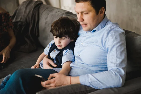 Father with little son sitting on the sofa. Father\'s day. Happy young father embracing kid sitting on sofa, loving caring dad hugging little boy. Father with little boy using digital tablet at home