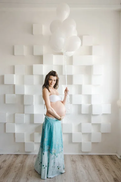 女人摸她怀孕的肚子一个美丽的孕妇在白色墙壁的背景的肖像。一个美丽的孕妇,有一个白色的球。怀孕的腹部和气球. — 图库照片