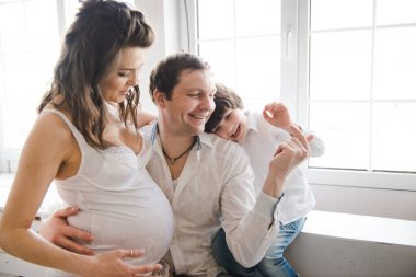 Mutlu aile ikinci bir çocuk bekliyor. Kocası ve küçük çocuğu olan hamile kadın. İkinci gebelik, annelik konsepti. Pencerenin yanında mutlu bir aile. Beyaz arka plan.