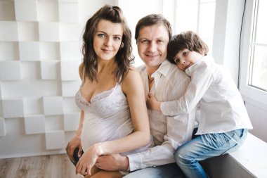 Mutlu aile ikinci bir çocuk bekliyor. Kocası ve küçük çocuğu olan hamile kadın. İkinci gebelik, annelik konsepti. Pencerenin yanında mutlu bir aile. Beyaz arka plan.
