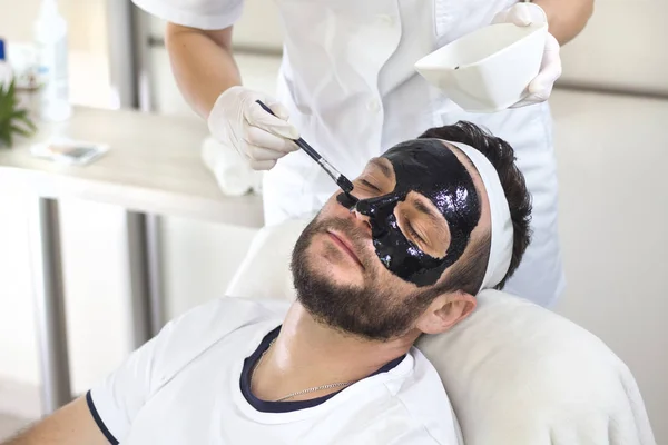 Beautician in a white dress and disposable gloves applies a black mask on the man\'s face.