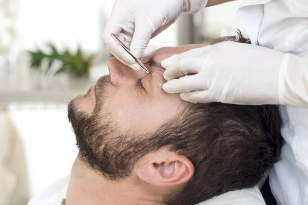 Beautician during the eyebrow adjustment procedure for a man. Removing unnecessary hair on eyebrows
