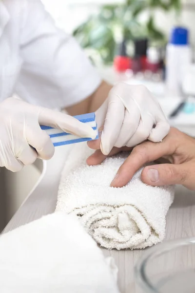Beautician in white disposable gloves in a beauty salon during a manicure treatment manicures his nails with a nail file in the palm of a man