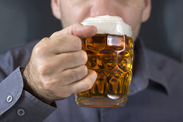 The man holds a beer mug in his hand with a light beer with foam on top and puts it in his mouth.