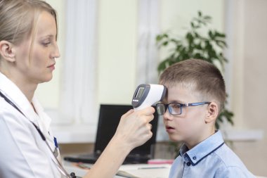Beyaz önlük ve boyun etrafında bir stetoskop Doktor doktor ofisinde oturuyor. Oğlan mavi tişört ve elektronik bir kızılötesi termometre kullanarak gözlük sıcaklığını ölçer