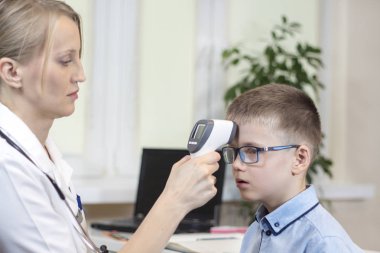 Doctor in a white coat and a stethoscope around the neck is sitting in the doctor's office. He measures the temperature of the boy in a blue shirt and glasses using an electronic infrared thermometer clipart