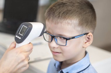 Doctor's hand measures the temperature of the boy in a blue shirt and glasses using an electronic infrared thermometer. clipart