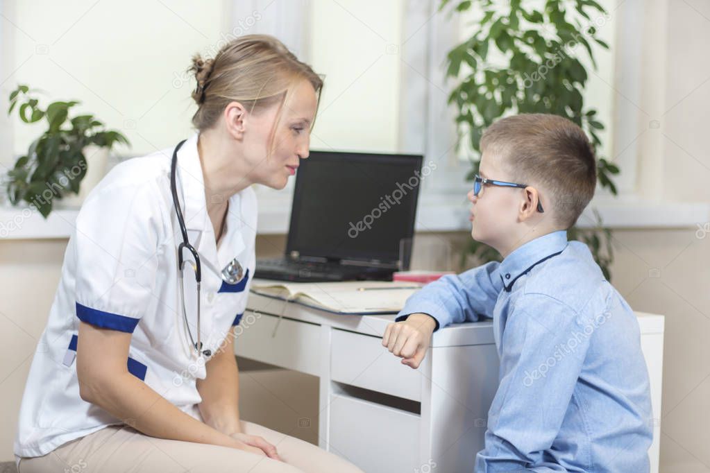 Doctor in a white coat and a stethoscope around the neck is sitting in the doctor's office. He talks to the boy in a blue shirt and glasses.
