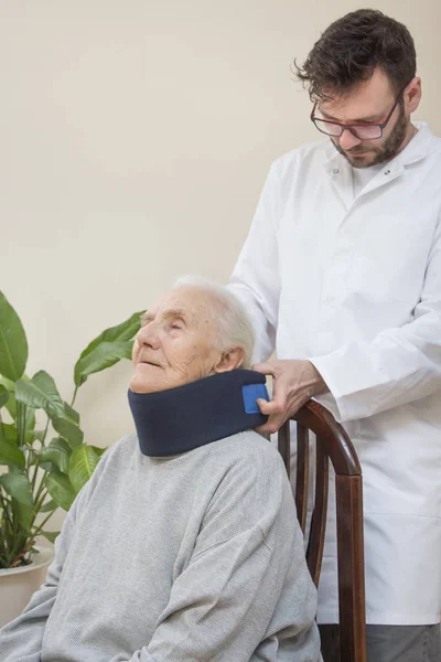 The orthopedic surgeon puts on a white orthopedic collar of a very old woman sitting on a chair.