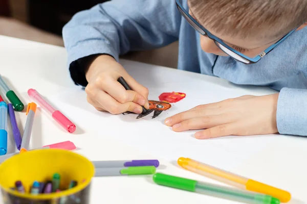 身穿蓝色衬衫和眼镜的男孩坐在一张白色的桌子前 用蜡笔在一张白纸上画画 在前景五颜六色的毛毡笔尖笔 — 图库照片