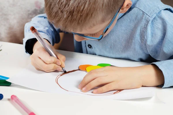 穿着蓝色衬衫和眼镜的男孩坐在办公桌前 专注于用彩纸做塑料工作 这个男孩正在用一支棕色的毛毡笔尖作画 — 图库照片