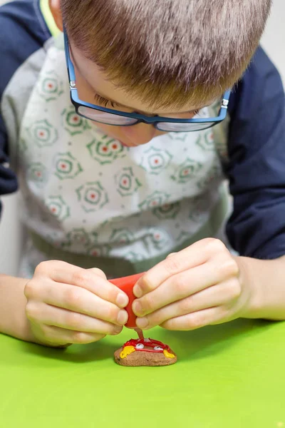 专注于男孩的脸工作 眼镜和厨房围裙 装饰饼干使用糖霜在管 装饰精美的蛋糕躺在前景的绿色桌子上 — 图库照片