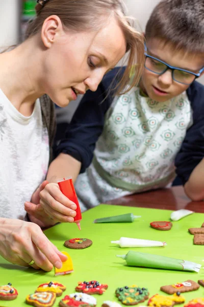 Mother and her son in the kitchen. A woman decorates a cinnamon cake with a tube of red icing. The son carefully observes what his mother is doing. Decorated cakes lie on a green table in the foreground.