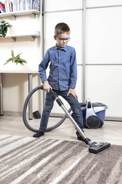 Cleaning an apartment by a child of school age. Children's home duties. The boy in glasses and a blue shirt is cleaning the flat. Vacuuming the apartment with a vacuum cleaner by a school-age boy.