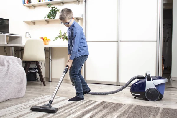 Cleaning an apartment by a child of school age. Children\'s home duties. Vacuuming a rug in an apartment by a boy dressed in a blue shirt and jeans pants.