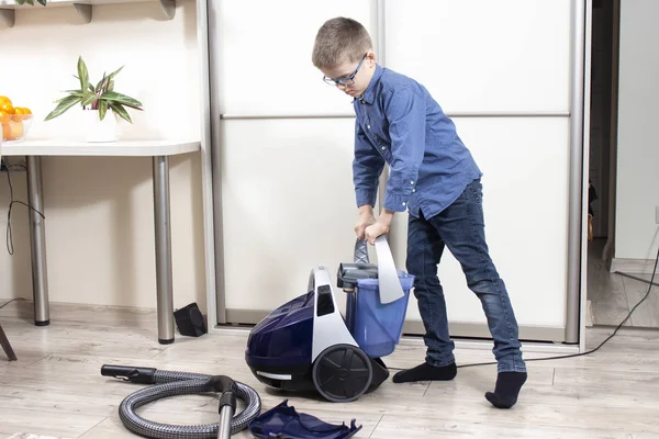 The boy in glasses and a blue shirt is cleaning the flat. Vacuuming the apartment with a vacuum cleaner by a school-age boy.