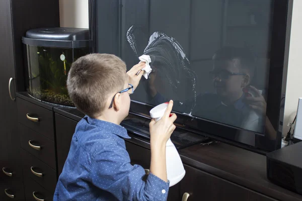 The school-age boy cleans the room, applies the detergent to the TV screen. In his hand he holds a white cloth to wipe away dust. Boy with glasses during cleaning. He wipes the TV screen with dust from the cloth.