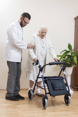 Beyaz bornozlu ve gecelikli yaşlı kadın rehabilitasyon yürüteciyle gider. Hemşire yaşlı kadını destekler ve korur..