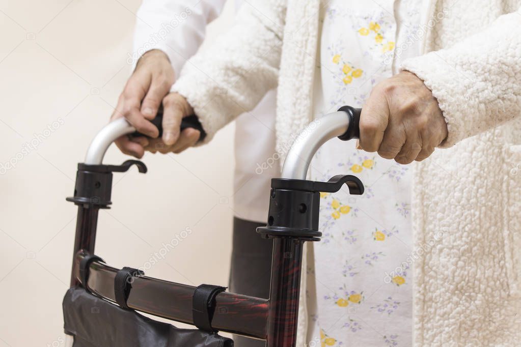 The hands of a very old woman hold the handles of a rehabilitation walker. The man nurse belay and helps in walking.