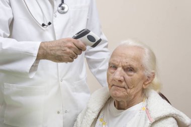 Boynunda stetoskop bulunan beyaz önlüklü sakallı doktor lazer termometreli çok yaşlı gri bir kadının sıcaklığını ölçüyor. Beyaz önlüklü doktor, çok yaşlı gri bir kadının lazer termometresi ile sıcaklığı ölçer..