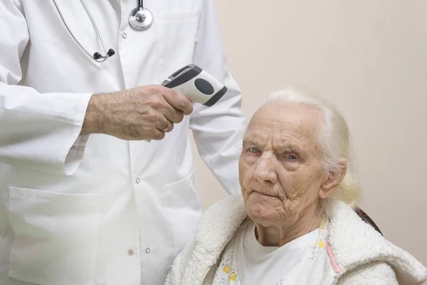 Bearded doctor in a white coat with a stethoscope around his neck measures the temperature of a very old gray woman with a laser thermometer. Doctor in a white coat measures the temperature with a laser thermometer of a very old gray woman.