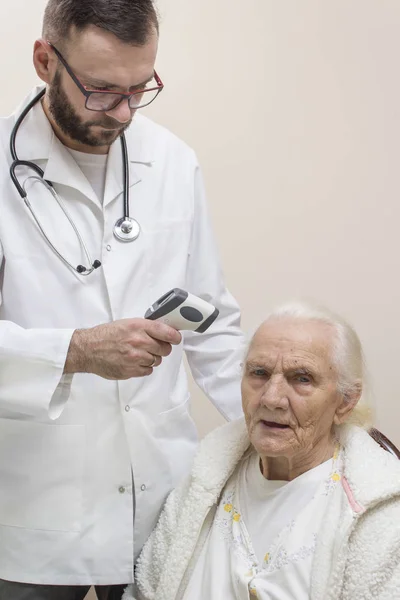 The doctor in a white coat measures the temperature with a laser thermometer of a very old gray woman. A bearded doctor in a white coat with a stethoscope around his neck measures the temperature of a very old gray woman with a laser thermometer.