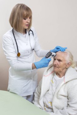 Tek kullanımlık eldivenli doktor yaşlı bir kadını muayene ediyor. Lazer termometre ile sıcaklığın ölçülmesi.