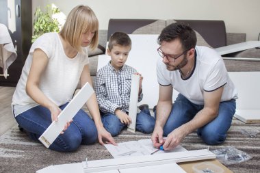 Aile bej bir halının üzerinde diz çöküyor. Önlerinde büyük bir özenle okudukları bir mobilya montaj kılavuzu var..