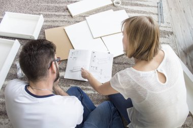 Kendi kendine montaj için beyaz mobilya elemanları odanın etrafına serilir. Kadın ve adam halının üzerinde oturuyorlar ve halıya yayılan talimatlara bakıyorlar..