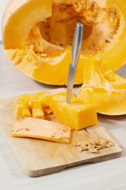 A kitchen knife stuck in a piece of pumpkin lying on a chopping board. The skin and pumpkin seeds lie on a wooden chopping board. A large fresh pumpkin cut in half lies on the kitchen counter.