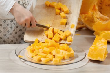 Mutfak önlüklü bir kadın doğrama tahtasından kesilmiş balkabaklarını cam bir tabağa aktarıyor..