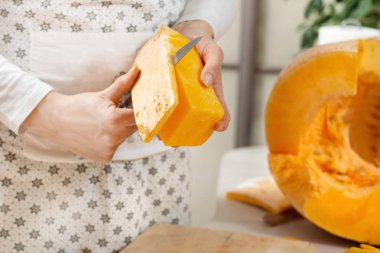 Dişi eller bir parça balkabağı tutuyor ve bir mutfak bıçağıyla derisini yüzüyor. Taze portakal kabağının yarısı mutfak tezgahında yatıyor..