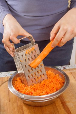 Dişi eller çiğ bir havucu tutar ve metal bir mutfak rendesinde küçük parçalara ayırır..