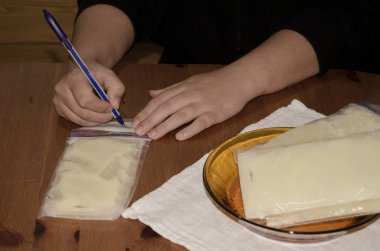 Anne dondurucuda saklamak için dondurulmuş anne sütünü torbaya koyuyor. Sütün ne zaman alındığını yazıyor. Sadece eller ve ahşap masa.