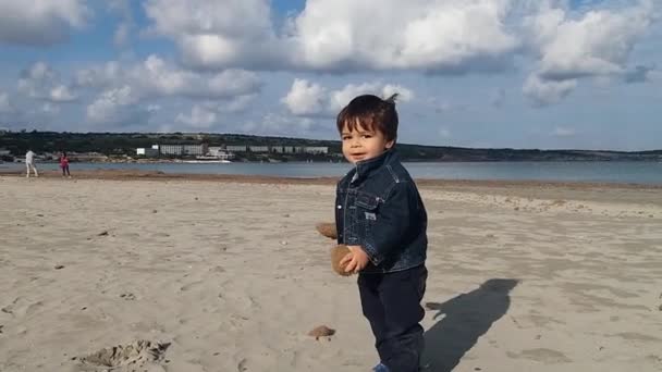 Eineinhalbjähriger Junge spielt mit Seegras-Bällen am Sandstrand ohne Menschen, sonniger Tag mit blauem Himmel und weißen Wolken — Stockvideo