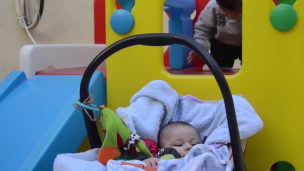 可爱的树月大的男婴享受在携带婴儿床 而他的哥哥是玩塑料玩房子滑梯 — 图库视频影像