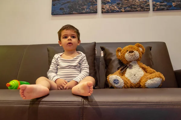 Lindo niño de dos años sentado en el sofá con su oso de peluche y viendo la televisión —  Fotos de Stock