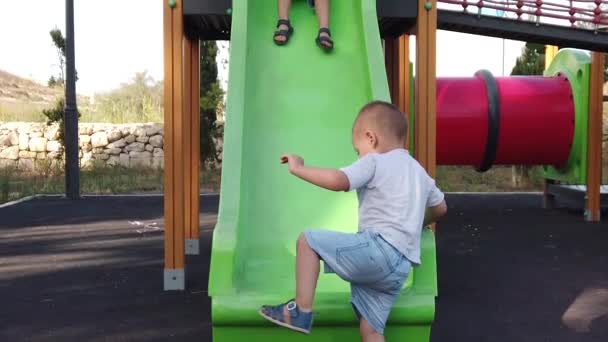 Schattige peuter klimt op de glijbaan terwijl zijn broer probeert naar beneden te glijden. Langzame beweging — Stockvideo