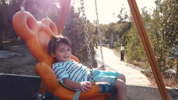 Adorabile ragazzo, bambino in età prescolare che oscilla sull'altalena in un parco vuoto e saluta alla telecamera al rallentatore — Video Stock