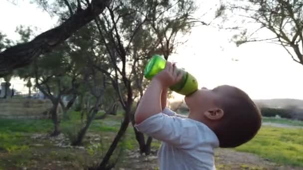 Schattige baby jongen drinkwater in het park tijdens zonsondergang — Stockvideo